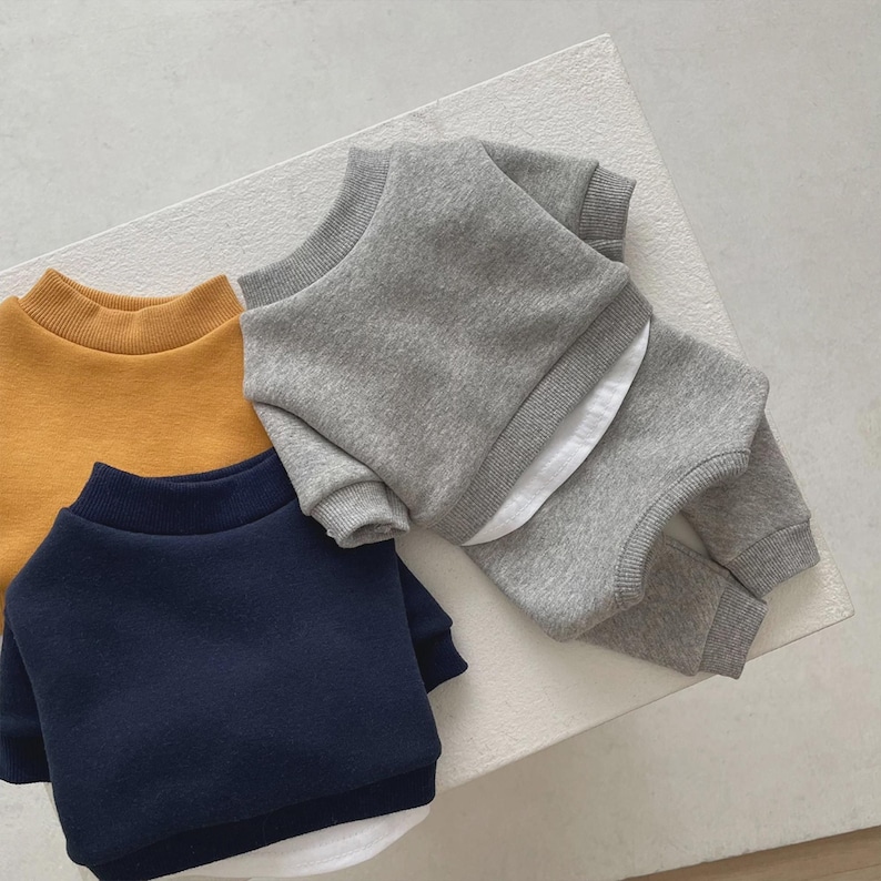 May include: Three children's sweatshirts in mustard yellow, navy blue, and gray. The gray sweatshirt is paired with matching pants. Each sweatshirt has a mock neck and a white undershirt peeking out from the bottom.