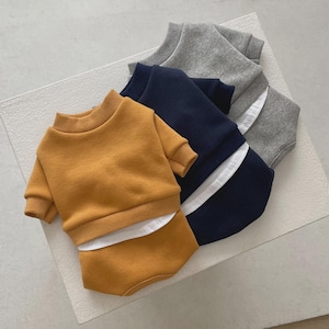 May include: A stack of baby outfits in mustard yellow, navy blue, and gray. Each set includes a short-sleeved sweatshirt and matching shorts. The navy blue and gray outfits have a white stripe detail. The outfits are made of a soft material.