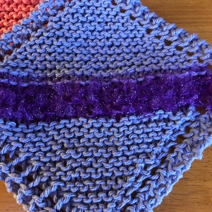 May include: A blue knitted dishcloth with a purple sparkly trim. The dishcloth is made with a simple knit stitch and has a lacy edge.