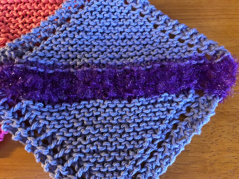 May include: A blue knitted dishcloth with a purple sparkly trim. The dishcloth is made with a simple knit stitch and has a lacy edge.