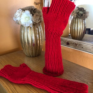 May include: A pair of red fingerless gloves, one is on a hand model and the other is lying flat on a wooden surface.