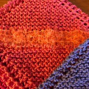 May include: Two knitted dishcloths, one in a pink and orange ombre pattern and the other in a solid purple. The pink and orange dishcloth has a strip of sparkly orange trim.