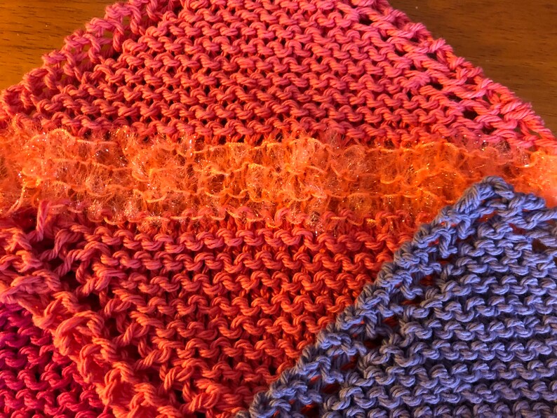 May include: Two knitted dishcloths, one in a pink and orange ombre pattern and the other in a solid purple. The pink and orange dishcloth has a strip of sparkly orange trim.