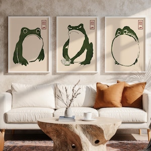 Könnte beinhalten: Drei gerahmte Kunstdrucke mit stilisierten grünen Frosch-Illustrationen auf beigem Hintergrund. Jeder Druck hat einen weißen Rahmen und ein rotes japanisches Zeichen. Die Drucke sind über einem weißen Sofa mit orangefarbenen Kissen ausgestellt.