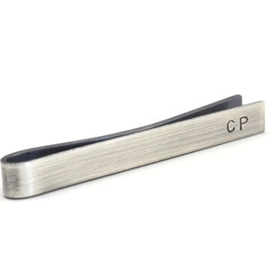 Darkened silver tie clip shown with slightly side-on view with dark edges of the tie clip and the unique finish from oxidising visible. Hand stamped initials are positioned towards the end on the front and are deep and dark showing up well.