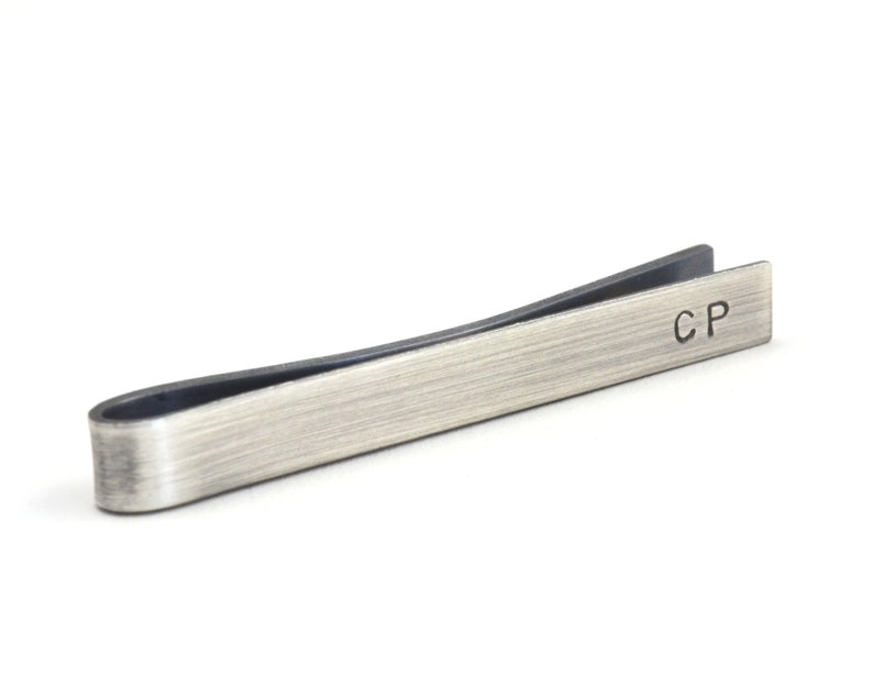 Darkened silver tie clip shown with slightly side-on view with dark edges of the tie clip and the unique finish from oxidising visible. Hand stamped initials are positioned towards the end on the front and are deep and dark showing up well.