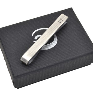 Oxidised Sterling silver tie clip shown resting on top of elegant black gift box. Box has simple logo in silver on front. This tie clip arrives as standard with this gift box ready for gifting. Tie clip is shown personalised with initials on front.