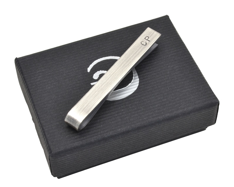 Oxidised Sterling silver tie clip shown resting on top of elegant black gift box. Box has simple logo in silver on front. This tie clip arrives as standard with this gift box ready for gifting. Tie clip is shown personalised with initials on front.
