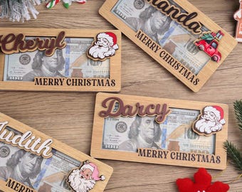 Könnte beinhalten: Weihnachtsgeschenkehalter aus Holz mit Namen wie "Amanda", "Cheryl", "Darcy" und "Judith". Jeder Halter zeigt ein Weihnachtsmann-Design und die Worte "Merry Christmas". Die Halter sind für eine US-$100-Rechnung konzipiert.