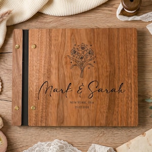May include: A rectangular wooden photo album with a dark brown wood grain finish. The cover features a black engraved floral bouquet, the names Mark & Sarah, the location New York, USA, and the date 25.02.2026. It has a black spine with gold screws.