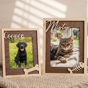 Op de afbeelding: Twee houten fotolijsten voor huisdieren. Eén lijst toont een zwarte hond genaamd "Cooper" met een botuitsparing. De andere lijst toont een kat genaamd "Misty" met een visgraatuitsparing. Beide lijsten hebben een lichte houten rand en een donkerder houten interieur.