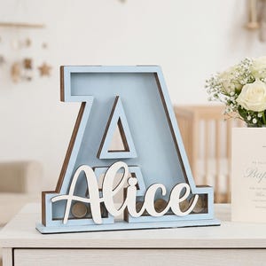 May include: A light blue wooden letter "A" shaped piggy bank with the name "Alice" in white script. The bank has a brown border and contains coins. A white card with "Baptism" text and a bouquet of white roses are also present.