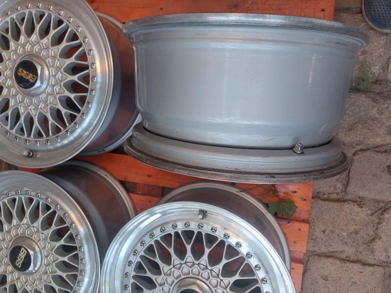 Puede incluir: Un juego de llantas de aleaci&oacute;n de color plateado con un dise&ntilde;o de m&uacute;ltiples radios. Las llantas tienen un acabado met&aacute;lico y est&aacute;n dispuestas sobre un palet de madera. Las tapas centrales muestran el logotipo "BBS".