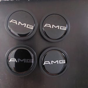 May include: Four black wheel center caps with the silver letters "AMG" in a bold, sans-serif font. The caps have a glossy finish and a circular design, likely for a car.