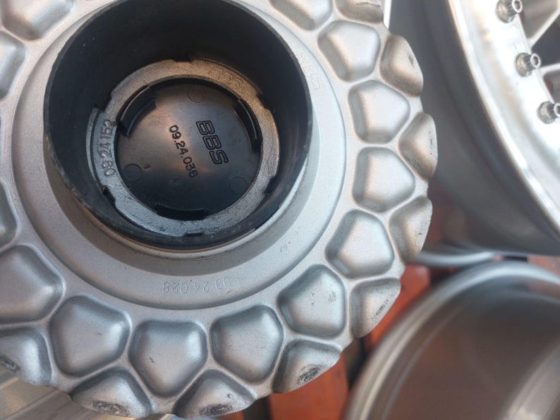 Puede incluir: Primer plano de un tapacubos plateado con un patr&oacute;n geom&eacute;trico. La tapa tiene el logotipo de BBS y los n&uacute;meros 09.24.036. La rueda es parcialmente visible en el fondo.