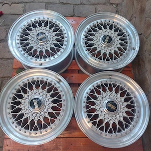 Puede incluir: Cuatro llantas de coche plateadas con un dise&ntilde;o de radios m&uacute;ltiples. Cada llanta tiene un buje central con el logotipo "BBS". Las llantas est&aacute;n dispuestas sobre una superficie de madera, mostrando sus intrincados patrones y acabado met&aacute;lico.