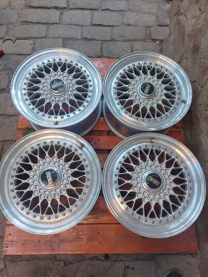 Puede incluir: Cuatro llantas de coche plateadas con un dise&ntilde;o de radios m&uacute;ltiples. Cada llanta tiene un buje central con el logotipo "BBS". Las llantas est&aacute;n dispuestas sobre una superficie de madera, mostrando sus intrincados patrones y acabado met&aacute;lico.