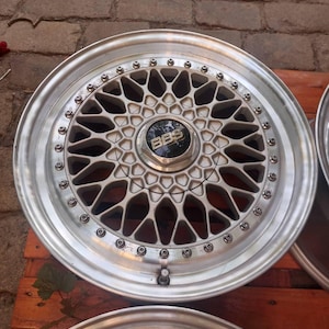 Puede incluir: Llanta de coche plateada con un dise&ntilde;o de radios geom&eacute;trico complejo. La tapa central presenta el logotipo "BBS" en negro. La llanta tiene un acabado met&aacute;lico pulido. El di&aacute;metro de la llanta es de aproximadamente 38 cm, para uso automotriz.