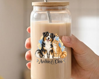 Può includere: Un bicchiere trasparente con coperchio in bambù e cannuccia in metallo, riempito con una bevanda beige. Il vetro presenta un'illustrazione colorata di un cane e un gatto con il testo Archie & Cleo stampato in un carattere corsivo scuro.