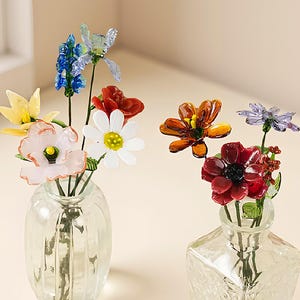 以下が含まれることがあります： カラフルなガラスの花のコレクションが入った2つの透明なガラスの花瓶。1つの花瓶は丸く、もう1つは四角です。花には赤、白、黄色、青、オレンジの花が含まれており、鮮やかなディスプレイを作成しています。