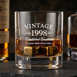 Puede incluir: Un vaso de cristal transparente lleno de líquido ámbar y dos cubos metálicos dorados. El vaso presenta un texto grabado en blanco que dice VINTAGE 1998, Established Excellence y CLASSIC BREW SINCE THEN, con dos estrellas flanqueando la última línea.