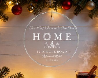 Pode incluir: Um enfeite de Natal circular transparente com o texto "Our First Christmas In Our New HOME" e o endereço "12 Dingle Road". O enfeite é decorado com desenhos de árvores de Natal e coroas. A data "5.20.2025" também está incluída.