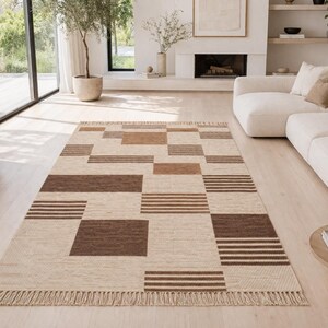 Könnte beinhalten: Ein rechteckiger Teppich mit einem geometrischen Muster in Beige- und Brauntönen. Der Teppich hat Quadrate und Streifen mit einem Fransenrand. Der Teppich liegt auf einem hellen Holzboden in einem Wohnzimmer.