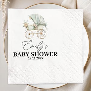 May include: A white square napkin with a baby carriage illustration in soft green and beige tones. The text "Emily's BABY SHOWER 19.11.2025" is printed in black script and block letters. The napkin is placed on a white plate with gold cutlery.