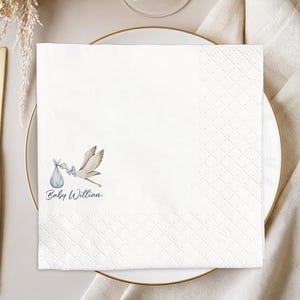 May include: White paper napkin with a stork carrying a baby in a blue bundle. The napkin is placed on a white plate with a gold rim. The text "Baby William" is printed in a script font below the stork illustration. Gold silverware is to the left.