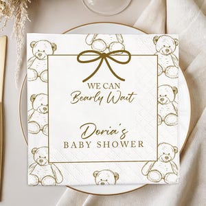 May include: A square white napkin with a gold border and teddy bear illustrations. The text "We Can Bearly Wait" and "Doria's Baby Shower" are printed in gold. The napkin is on a white plate with gold silverware.