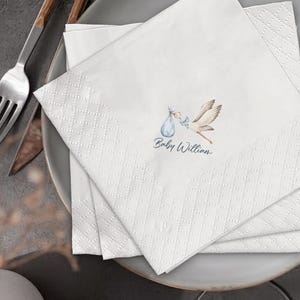May include: White paper napkins with a diamond pattern. Each napkin features a stork carrying a baby bundle, with the text "Baby William" in blue script. The napkins are stacked on a gray plate.