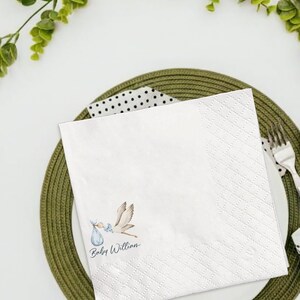 May include: White square paper napkin with a diamond pattern and a stork carrying a baby in a blue bundle. The text "Baby Willian" is printed in a script font. The napkin is on a green plate with a fork.