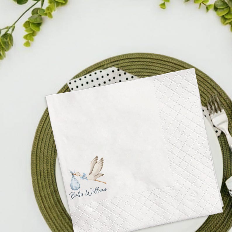 May include: White square paper napkin with a diamond pattern and a stork carrying a baby in a blue bundle. The text "Baby Willian" is printed in a script font. The napkin is on a green plate with a fork.
