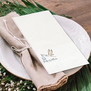 May include: White paper napkin with a watercolor illustration of a stork carrying a baby in a blue bundle. The text "Baby Willian" is written in a delicate script below the stork. The napkin is placed on a white plate with a beige linen napkin.