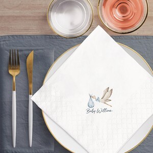 May include: White paper napkin with a stork carrying a baby in a blue bundle. The napkin is on a white plate with gold trim, and the text "Baby William" is printed on the napkin. Gold and white silverware is to the left.