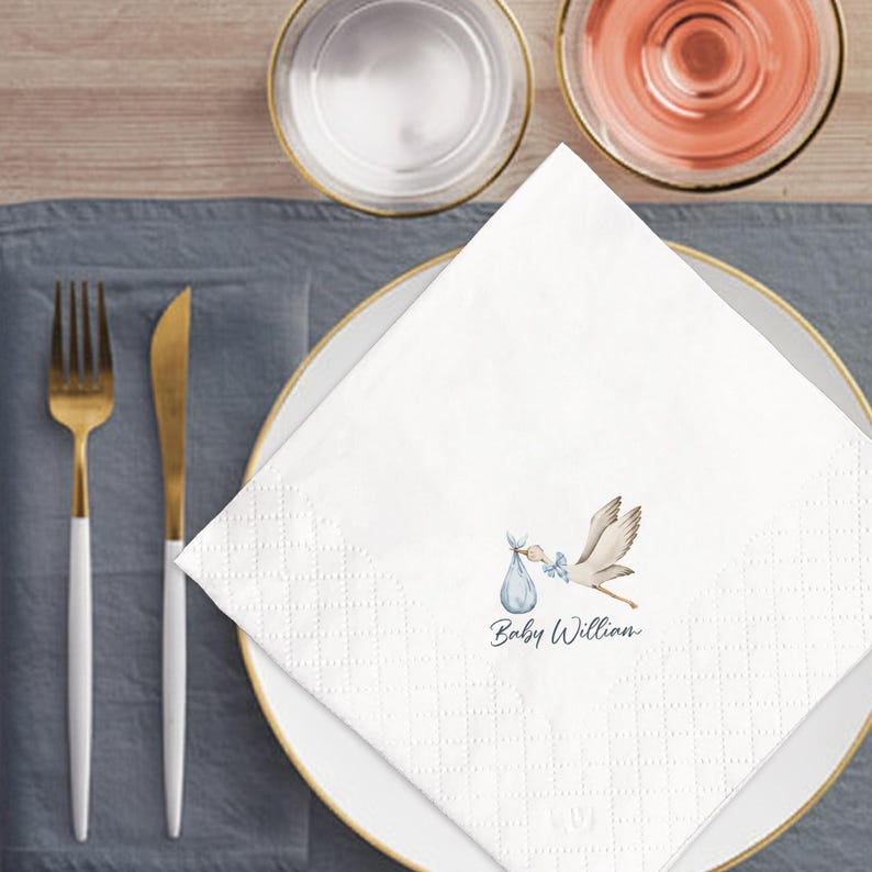 May include: White paper napkin with a stork carrying a baby in a blue bundle. The napkin is on a white plate with gold trim, and the text "Baby William" is printed on the napkin. Gold and white silverware is to the left.