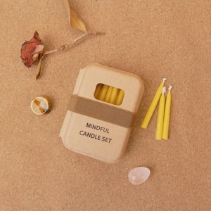 Könnte beinhalten: Ein beigefarbenes Box-Set mit der Aufschrift "MINDFUL CANDLE SET" und einem braunen Band, das gelbe Kerzen enthält. Drei weitere gelbe Kerzen befinden sich neben der Box. Eine getrocknete Rose, ein kleines Dekorationsobjekt und ein klarer ovaler Stein sind ebenfalls vorhanden.