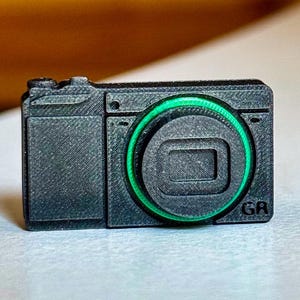 Op de afbeelding: Een kleine, zwarte camera met een groene lensring en de letters "GR" op de voorkant. De camera staat op een wit oppervlak.
