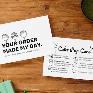 Könnte beinhalten: Zwei weiße Karten mit schwarzem Text auf einer Holzoberfläche. Auf einer Karte steht "YOUR ORDER MADE MY DAY" mit Cake-Pop-Illustrationen. Die andere Karte sagt "Cake Pop Care" mit Anweisungen. Ein grüner Bleistift und ein Notizbuch sind ebenfalls sichtbar.