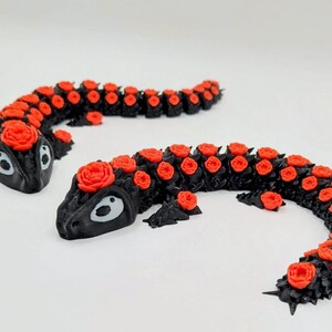 Könnte beinhalten: Zwei schwarze, stilisierte Salamander-Skulpturen mit roten Rosenakzenten. Die Salamander haben große, ovale Augen und sind auf einer weißen Oberfläche platziert. Die Rosen sind gleichmäßig auf dem Rücken der Salamander verteilt.
