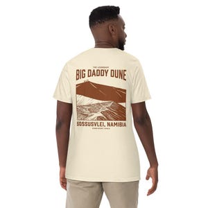 Peut inclure: Un t-shirt à manches courtes de couleur crème avec un motif marron au dos. Le design représente un paysage de dunes désertiques avec le texte THE LEGENDARY BIG DADDY DUNE, SOSSUSVLEI, NAMIBIA et NAMIB DESERT, AFRICA imprimé en lettres marron.