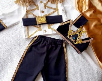May include: A white and navy blue prince costume set with gold detailing. The set features a jacket with gold fringe and trim, navy blue trousers with gold trim, and black shoes with gold embellishments. A gold satin cape is also included.