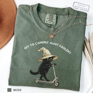 May include: Moss green t-shirt with the phrase "OFF TO COMMIT AUNT FOOLERY" and a graphic of a black cat wearing a witch hat and riding a scooter. A gold necklace and a brown hat are also visible.