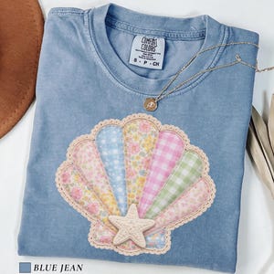 May include: Blue jean Comfort Colors t-shirt with a seashell design. The seashell is made of various patterned fabrics, including floral and gingham, and features a starfish. A gold necklace rests on the shirt.