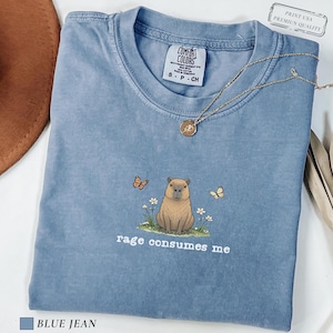 Blue Jean Comfort Colors® t-shirt featuring a vintage capybara illustration sitting in grass with small flowers and butterflies, with the phrase “rage consumes me”.
