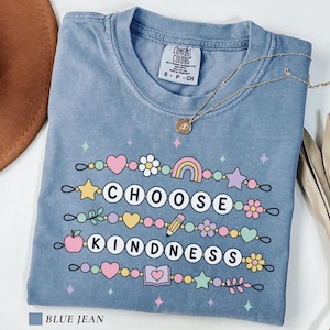 May include: A blue jean colored t-shirt featuring a graphic print of beaded strings with the text CHOOSE KINDNESS. The design includes colorful icons like hearts, stars, flowers, a rainbow, a pencil, an apple, and a book, with small sparkles.