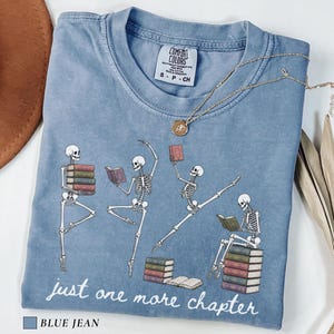 May include: A blue jean colored t-shirt featuring a graphic of four skeletons reading and dancing with stacks of books. The text "just one more chapter" is printed in white below the skeletons. The shirt has a crew neckline and a Comfort Colors label.