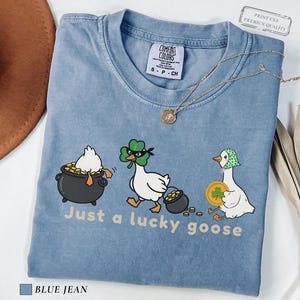 blue jean comfort colors t-shirt with three illustrated geese, one in a pot of gold, one wearing a shamrock mask, and one holding a coin, playful st patrick’s day design