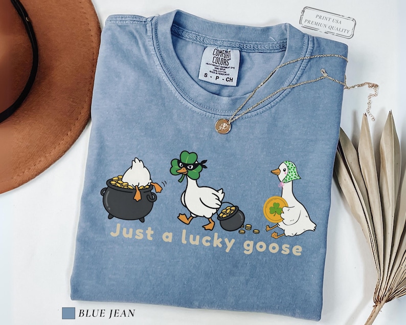 blue jean comfort colors t-shirt with three illustrated geese, one in a pot of gold, one wearing a shamrock mask, and one holding a coin, playful st patrick’s day design
