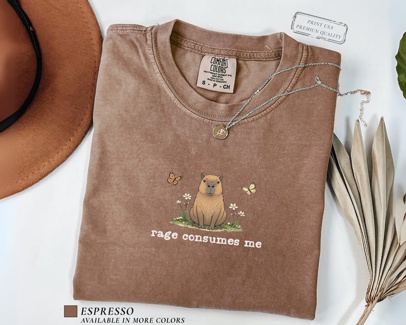 Espresso Comfort Colors® t-shirt featuring a vintage capybara illustration sitting in grass with small flowers and butterflies, with the phrase “rage consumes me”.